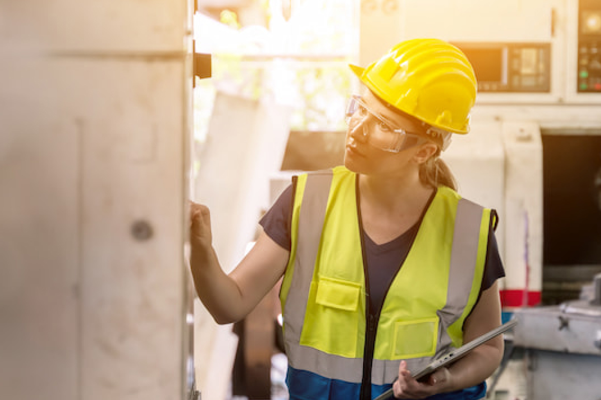 Woman in yellow hardhat and vest inspecting manufacturing equipment and taking notes